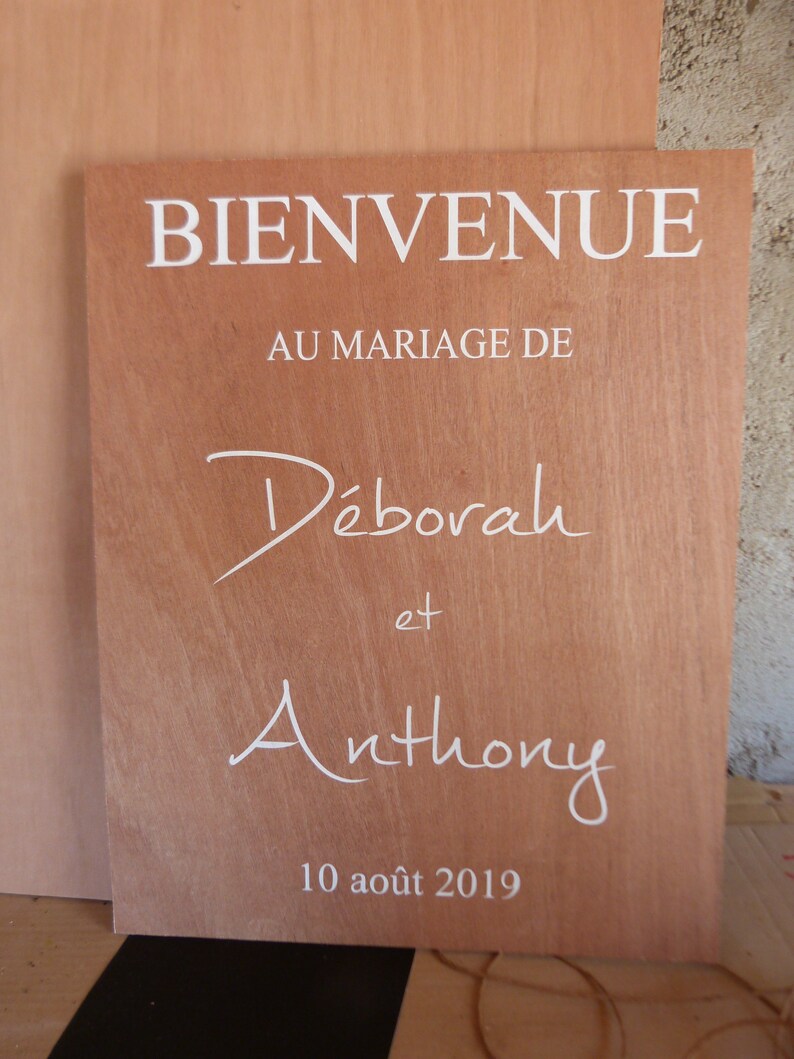 May include: A wooden sign with the text "BIENVENUE AU MARIAGE DE D&eacute;borah et Anthony 10 ao&ucirc;t 2019" in white lettering.