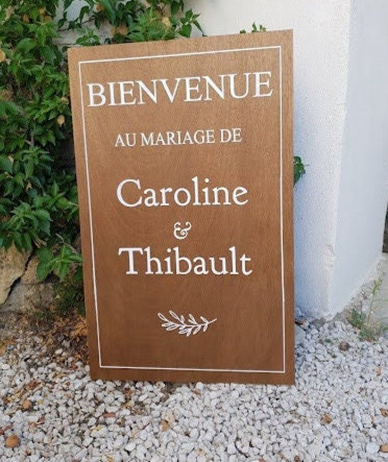 May include: A brown wooden sign with white text that reads "Bienvenue au mariage de Caroline & Thibault" with a small branch design at the bottom.