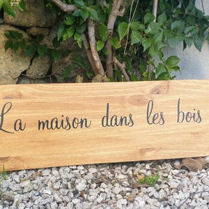 Può includere: Un cartello di legno con il testo "La maison dans les bois" in lettere nere.