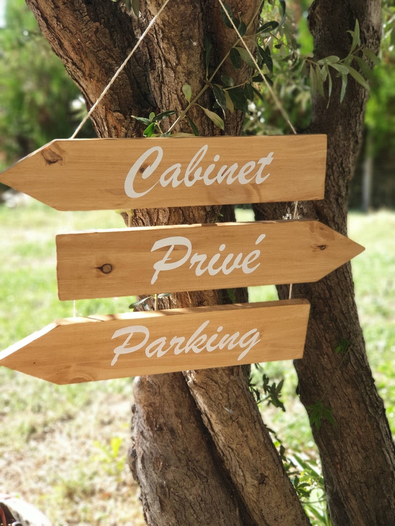 May include: Three wooden arrow signs hanging from a tree branch. The signs point to the right and are labeled "Cabinet", "Priv&eacute;", and "Parking" in white cursive lettering.