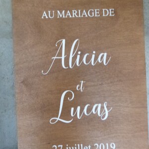 May include: A wooden sign with the text "BIENVENUE AU MARIAGE DE Alicia et Lucas 27 juillet 2019" in white lettering.