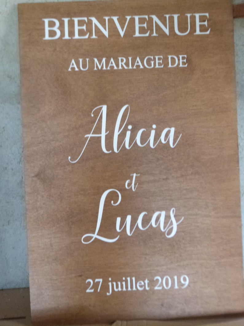 May include: A wooden sign with the text "BIENVENUE AU MARIAGE DE Alicia et Lucas 27 juillet 2019" in white lettering.