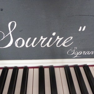 May include: A white digital piano with black keys. The top of the piano has a gray surface with the words "Sourire" and "Soprano" written in white.
