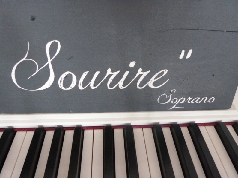 May include: A white digital piano with black keys. The top of the piano has a gray surface with the words "Sourire" and "Soprano" written in white.