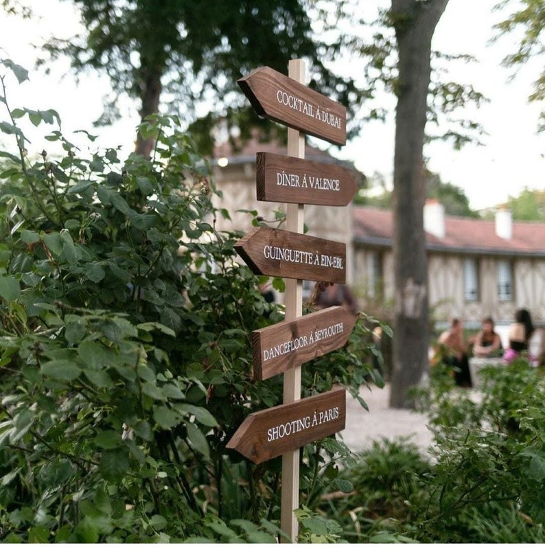 May include: Wooden signpost with five arrows pointing in different directions. Each arrow has a white text label: Cocktail &agrave; Dubai, Diner &agrave; Valence, Guinguette &agrave; Ein-Erl, Dancefloor &agrave; Beyrouth, Shooting &agrave; Paris.