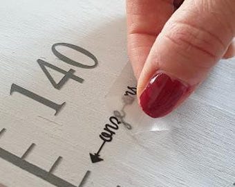 May include: A white wooden growth chart with a gray ruler and black markings. The number 140 is visible on the chart. A clear sticker with the words "one year" and an arrow pointing to the right is being applied to the chart.