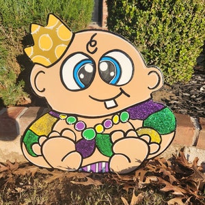 May include: A cartoon baby wearing a yellow crown with polka dots, a purple and green beaded necklace, and a purple, green, and gold costume. The baby has large blue eyes and a toothy grin.