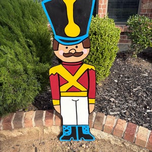 May include: A wooden toy soldier yard decoration. The soldier has a black hat with a yellow accent, a red jacket with yellow cross straps, white pants, and blue boots. The soldier has a brown mustache and is standing upright.