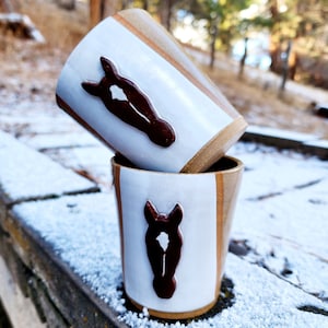 May include: Two white ceramic mugs with brown horse head designs. The mugs have a brown and white striped pattern. The mugs are sitting on a wooden surface covered in snow.