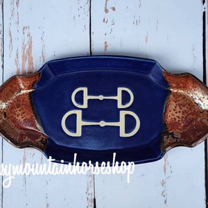 May include: A blue ceramic dish with a white design of two horse bits. The dish has a brown rim and is made in Montana, USA. @snowymountainhorseshop