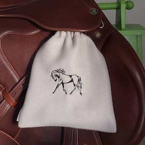 May include: A white fleece saddle bag with a black embroidered horse design. The bag is hanging on a brown leather saddle.