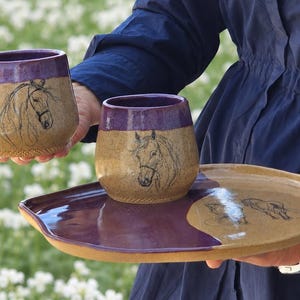 May include: A ceramic drinkware set featuring two stemless wine glasses and a serving tray. The glasses and tray have a tan base with a purple rim and accents. Each piece is decorated with a black horse head illustration.