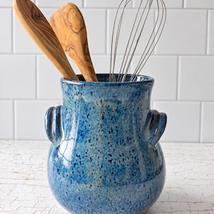 May include: A blue ceramic utensil crock with a speckled glaze and two side handles, containing two wooden spoons and a metal wire whisk. The vessel has a rounded body and a wide opening, showcasing a vibrant blue finish with dark spots.