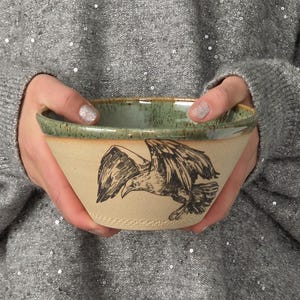 May include: A ceramic bowl with a beige exterior featuring a black ink illustration of a bird in flight. The bowl has a speckled sage green glazed rim and interior, showcasing a hand-thrown pottery aesthetic with a smooth, matte finish.