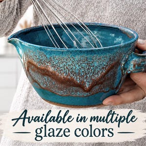 May include: A ceramic mixing bowl with a vibrant teal and brown reactive glaze, featuring a pouring spout and a side handle. A metal wire whisk is positioned inside the bowl. The text "Available in multiple glaze colors" is visible.