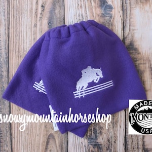 Puede incluir: Dos cubrebotas de forro polar morado con un diseño bordado en blanco de un caballo saltando una valla. El diseño también incluye el texto "Made in Montana USA".