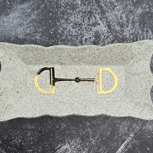 May include: A gray, rectangular tray with a scalloped edge holds a gold-colored horse bit. The bit is centered on the tray, with the D-shaped rings and the central bar clearly visible. The tray has a speckled texture.