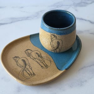 May include: A ceramic cup and matching oval plate featuring a speckled tan and blue glaze. The cup displays a black line drawing of a horse, while the plate features two similar horse illustrations. The pieces have a smooth, glossy finish.