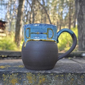 May include: A handmade ceramic mug with a two-tone design. The top half is a glossy blue with a gold horse bit design, and the bottom half is a textured dark brown. The handle is blue. The mug is sitting on a wooden surface.