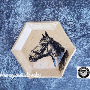 May include: A hexagonal ceramic dish with a speckled glaze in beige and white. The dish features a black and white silhouette of a horse's head with a bridle. The dish has a raised rim and a slightly textured surface. The text "@snowymountainhorseshop" is printed on the bottom of the dish. A round sticker with the text "Made in Montana USA" is visible in the background.