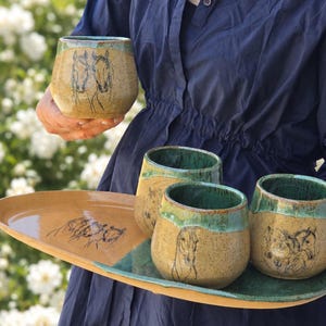 May include: A handmade ceramic drinkware set with horse illustrations. The set includes four stemless cups and a serving tray. The cups have a speckled tan body with a green rim and horse drawings. The tray is tan with a green accent.