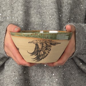 May include: A ceramic bowl with a matte beige exterior featuring a black ink illustration of a flying raven. The rim of the bowl is finished with a glossy, mottled green glaze that drips slightly down the sides of the textured clay surface.