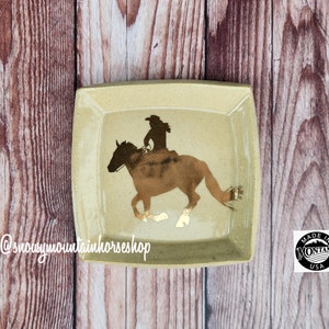 May include: A square, beige ceramic dish with rounded corners. The dish features a brown silhouette of a person riding a horse, with gold accents. The dish is marked "Made in Montana USA" and has the text "@snowymountainhorseshop".