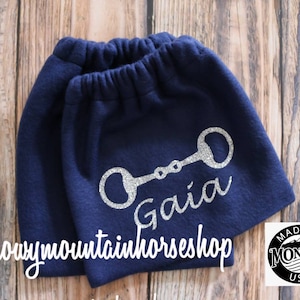 May include: A pair of navy blue fleece leg warmers with a silver glitter horse bit and the name "Gaia" written below it. The leg warmers are made in Montana, USA.