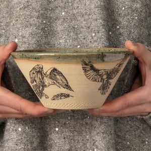 May include: A ceramic bowl with a light beige matte finish featuring black ink illustrations of two ravens in flight and a single feather. The bowl has a wide, flared rim with a speckled green and brown glaze, and a textured base.