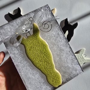 May include: A lime green ceramic horse ornament with a textured surface is presented in a silver gift box with a silver ribbon. Additional horse ornaments in black and white are visible. The ornament is a decorative item.