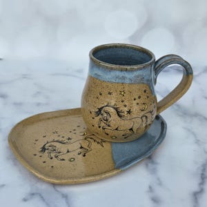 Puede incluir: Un juego de taza y plato de cerámica. La taza y el plato son de color beige con borde y base azules. El conjunto presenta una ilustración negra de un caballo, estrellas y una luna creciente. La taza tiene un asa curva.