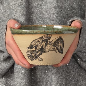 May include: A ceramic bowl with a light beige speckled finish featuring a black ink illustration of a bird of prey in flight. The bowl has a wide, flared rim glazed in a mottled sage green with brown accents along the top edge.