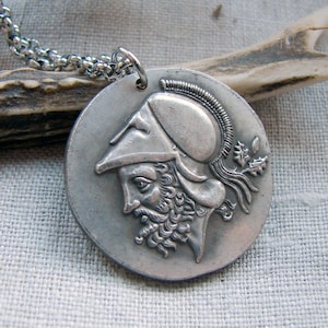 May include: A silver-toned pendant necklace featuring a detailed profile of a man in a helmet and beard. The round pendant is attached to a silver chain. The necklace is displayed on a neutral linen surface.
