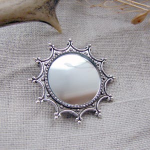 Peut inclure: Une broche circulaire de couleur argentée dotée d'un centre miroir réfléchissant, entouré d'un cadre orné de style soleil avec des détails perlés et des extensions pointues en forme d'arche rayonnant depuis le miroir central.
