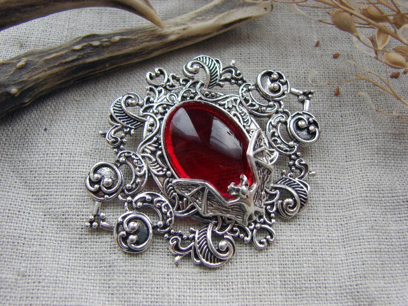 May include: A silver-toned metal brooch featuring an intricate filigree frame with swirling patterns. A large, oval-shaped, deep red faceted gemstone is set in the center, accented by a small, detailed silver bat figure perched at the bottom edge.