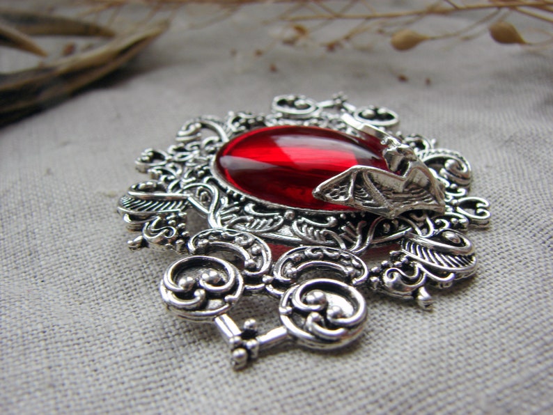 May include: A silver-toned brooch featuring an intricate filigree metal frame with a large, smooth, oval-shaped red gemstone in the center. A small, detailed metal bat figure is attached to the right side of the frame, resting over the red stone.