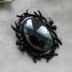 May include: A black brooch featuring a central oval-shaped polished stone with a reflective surface. The stone is encased in a dark, glossy frame designed to resemble a cluster of intertwined branches or thorns, creating a textured border.