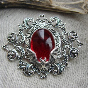 May include: A silver-toned brooch featuring an oval-shaped, deep red translucent cabochon at the center. The metal frame is decorated with intricate scrollwork and filigree patterns, with a small bat figure positioned at the bottom of the stone.