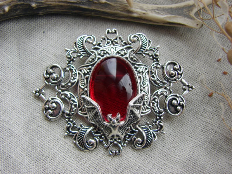 May include: A silver-toned brooch featuring an oval-shaped, deep red translucent cabochon at the center. The metal frame is decorated with intricate scrollwork and filigree patterns, with a small bat figure positioned at the bottom of the stone.
