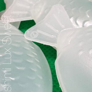 May include: Close-up of several translucent, fish-shaped molds. The molds are white and have a textured surface. The text "Sashimi Lux Sweet" is visible on the left side of the image.