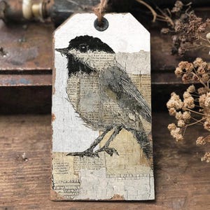 May include: A rectangular, white tag with a black and gray bird illustration. The bird is set against a background of vintage paper and text. A brown string is attached at the top. The tag has a rustic, distressed appearance.