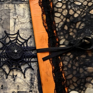 May include: A close-up of a handmade journal with a black lace overlay and a black ribbon tied around it. The journal has a textured, orange cover and a black spiderweb design on the front.