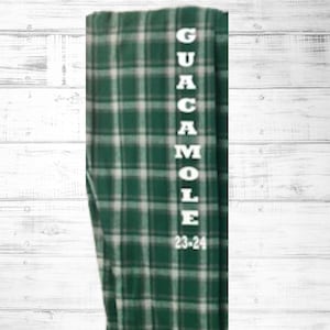 Pode incluir: Calças de pijama xadrez verdes e brancas com a palavra "GUACAMOLE" impressa verticalmente em branco. O número "23-24" está impresso abaixo da palavra "E". As calças estão dobradas e exibidas em um fundo de madeira branca.