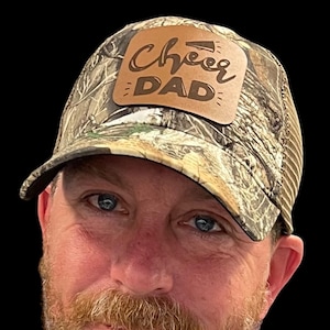 May include: A camouflage trucker hat with a brown leather patch that says "Cheer Dad" in a cursive font.