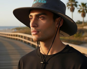 May include: A charcoal gray wide-brimmed sun hat featuring the text SUNSET BEACH embroidered in light blue on the front. The hat includes a black adjustable chin strap with a sliding toggle, ventilation eyelets, and a structured crown.