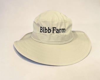 May include: A light beige bucket hat with a wide brim and a rounded crown. The front of the hat features the text Bibb Farm embroidered in black thread. The hat is constructed from a solid fabric with visible stitching along the brim and crown.