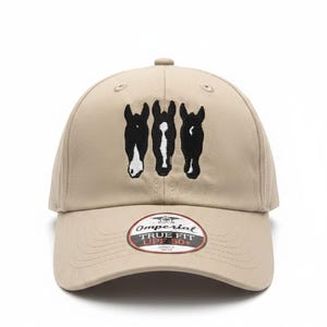 May include: A tan baseball cap featuring an embroidered design of three black horse heads with white markings. A circular sticker on the brim displays the text Omperial TRUE FIT UPF 50+ in black, white, and red colors against a tan background.