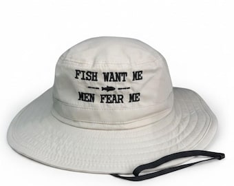 May include: A beige bucket hat featuring black embroidered text that reads FISH WANT ME with a small fish icon in the centre and MEN FEAR ME below it. The hat includes a wide brim and a thin black adjustable chin strap attached to the side.