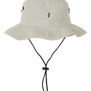 Pode incluir: Um chap&eacute;u de balde bege com um cord&atilde;o de ajuste preto e um bot&atilde;o preto. O chap&eacute;u tem uma aba larga e uma coroa plana.