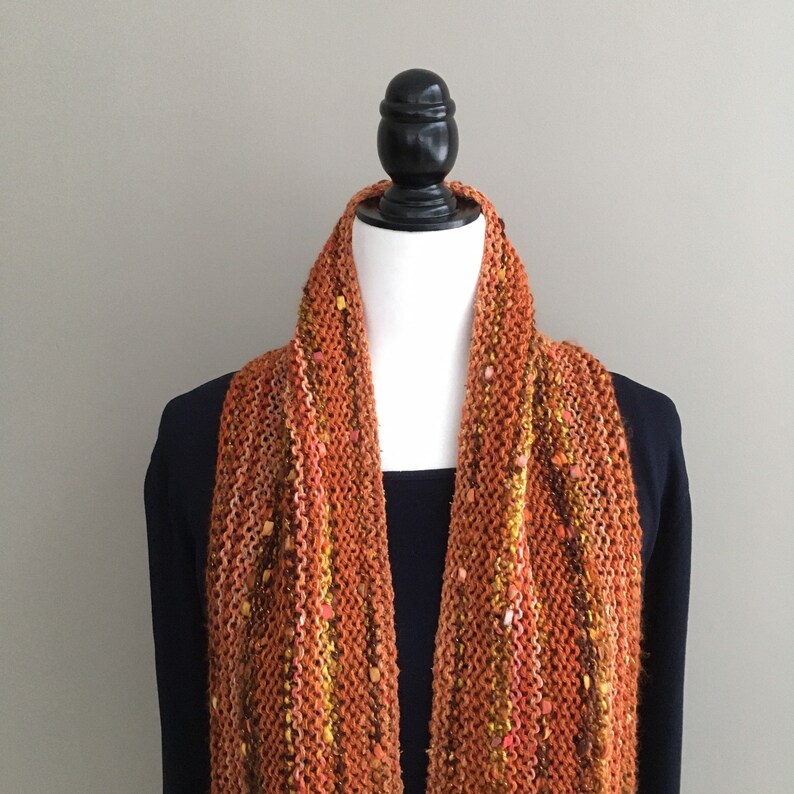 K&ouml;nnte beinhalten: Ein gestrickter Schal in Orange- und Braunt&ouml;nen mit gelben und rosa Tupfen. Der Schal wird auf einer Schaufensterpuppe pr&auml;sentiert. Ein warmes, strukturiertes Accessoire.